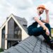 happy handyman in orange helmet sitting on roof and holding hammer